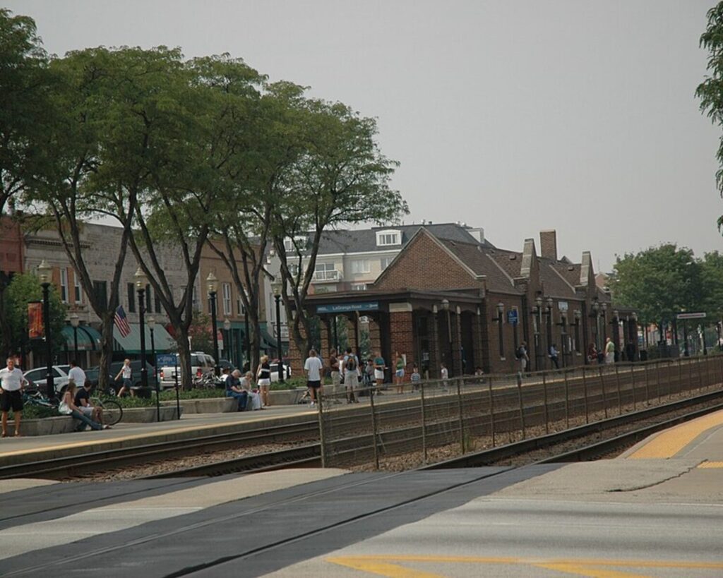 Train platform with tracks in the foreground, people waiting and sitting near benches, and a row of brick station buildings with trees behind them.