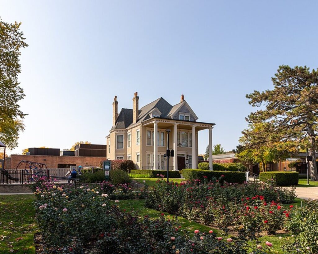 Two-story beige house with a columned porch, surrounded by blooming rose bushes and a manicured lawn under a clear blue sky.
