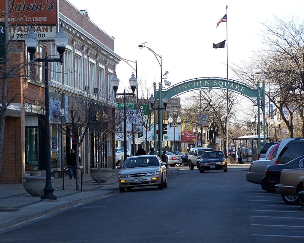 Lincoln Square commercial street with shops, parked cars, decorative lamps, and a neighborhood arch spanning the roadway.