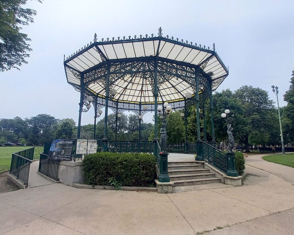 Decorative wrought-iron gazebo in a park with lamp posts and stairs leading up to a fenced platform.