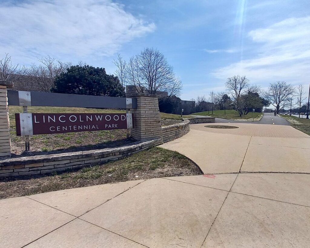 Entrance sign reading Lincolnwood Centennial Park beside a brick and metal frame, with a paved walkway extending into a grassy park under a blue sky.
