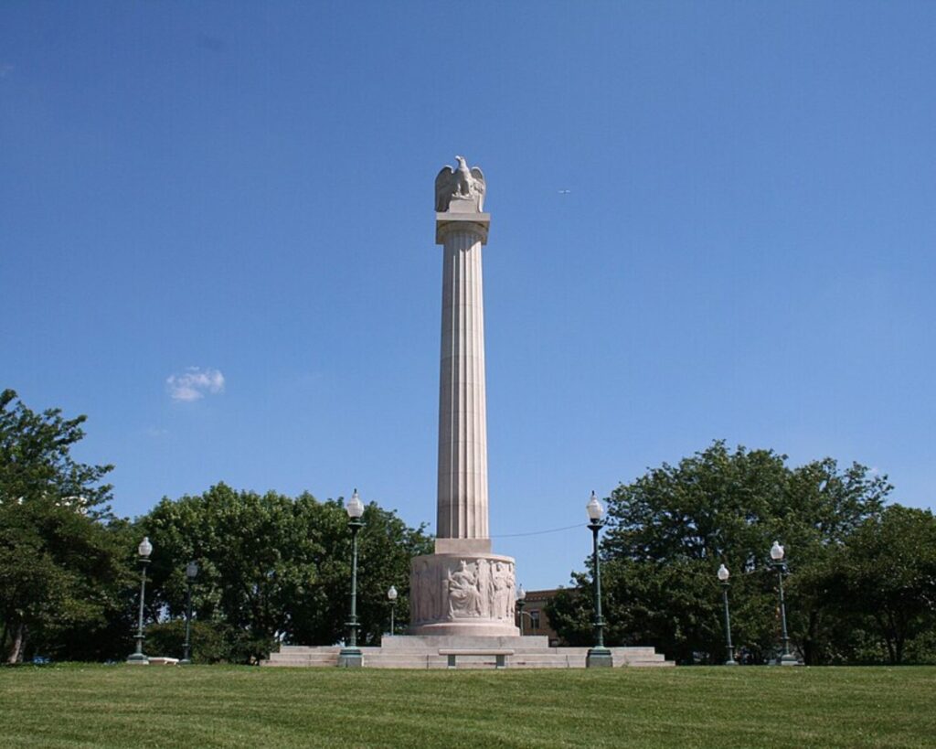 Tall stone column monument topped with an eagle statue in a grassy park, with trees and lampposts surrounding it