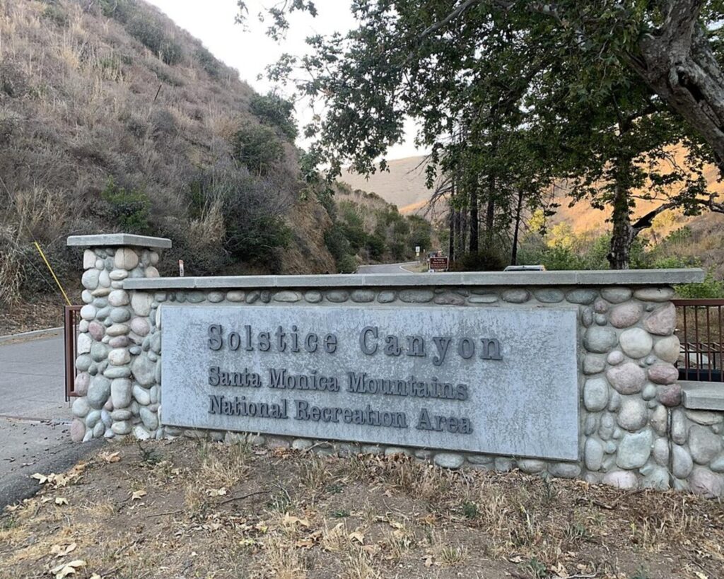 Stone entrance sign reading 'Solstice Canyon, Santa Monica Mountains National Recreation Area' with a stone wall and hillside backdrop.