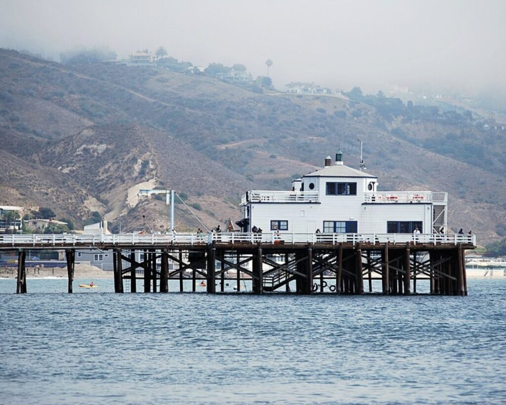 Malibu Pier from Surfrider Beach with ocean water, wooden pier, white building, and foggy hills.