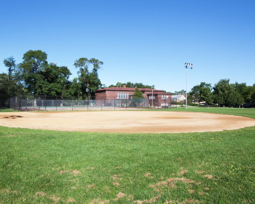 Baseball field at Rutherford Sayre Park in Chicago’s Montclare neighborhood, with a dirt infield, green lawn, fencing, trees, and a brick fieldhouse under a clear blue sky.