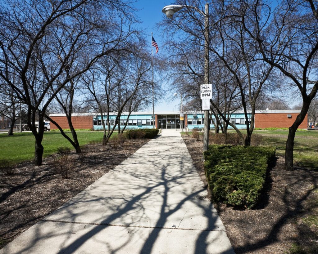 Kennedy Park fieldhouse in Chicago, viewed along a paved walkway through leafless trees with an American flag flying outside.