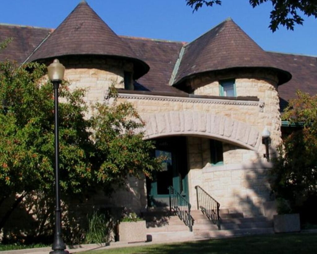 Stone entrance to the Walker Library with twin round turrets, conical roofs, an arched doorway, and front steps surrounded by trees.