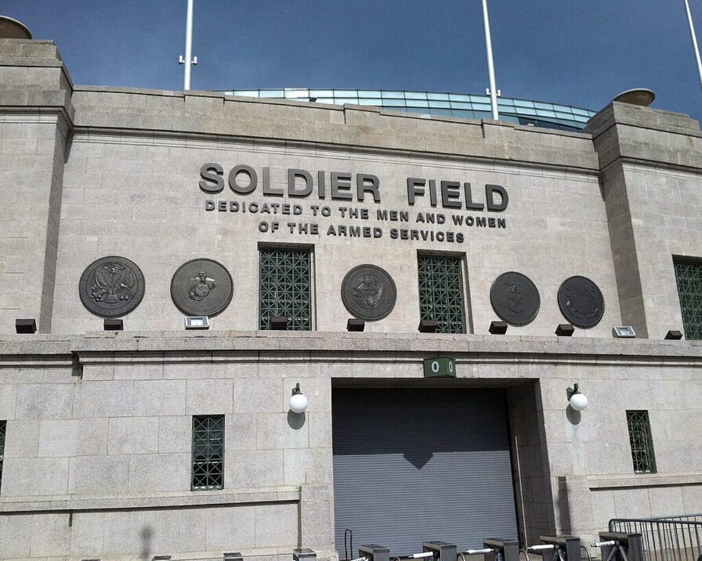 Exterior view of Soldier Field with large lettering: 'SOLDIER FIELD' and dedication to the men and women of the armed services, plus circular medals on the wall facade at ground level.