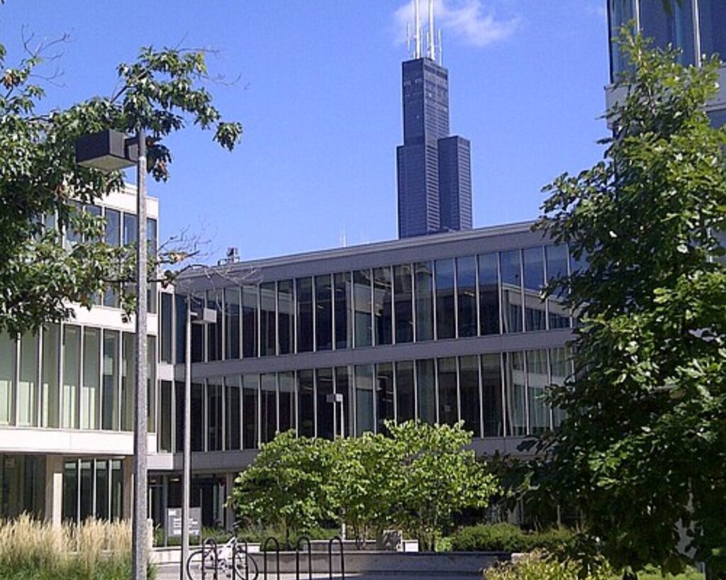 Modern glass-office building with a tall dark tower in the background, framed by green trees and a bike rack in the foreground.