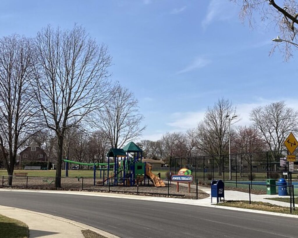 Playground with a blue and green play structure and slide inside a fenced park near a street on a sunny day.