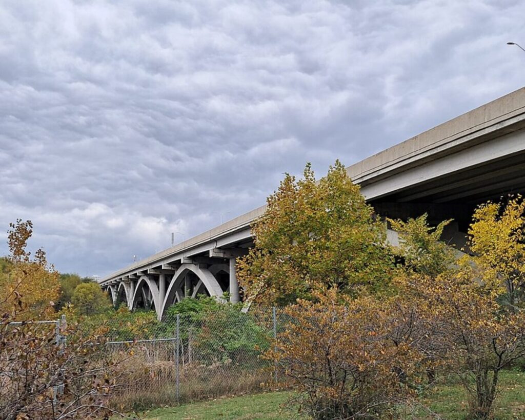 Concrete bridge with arched supports extending over a fenced, overgrown lot beneath a cloudy sky.
