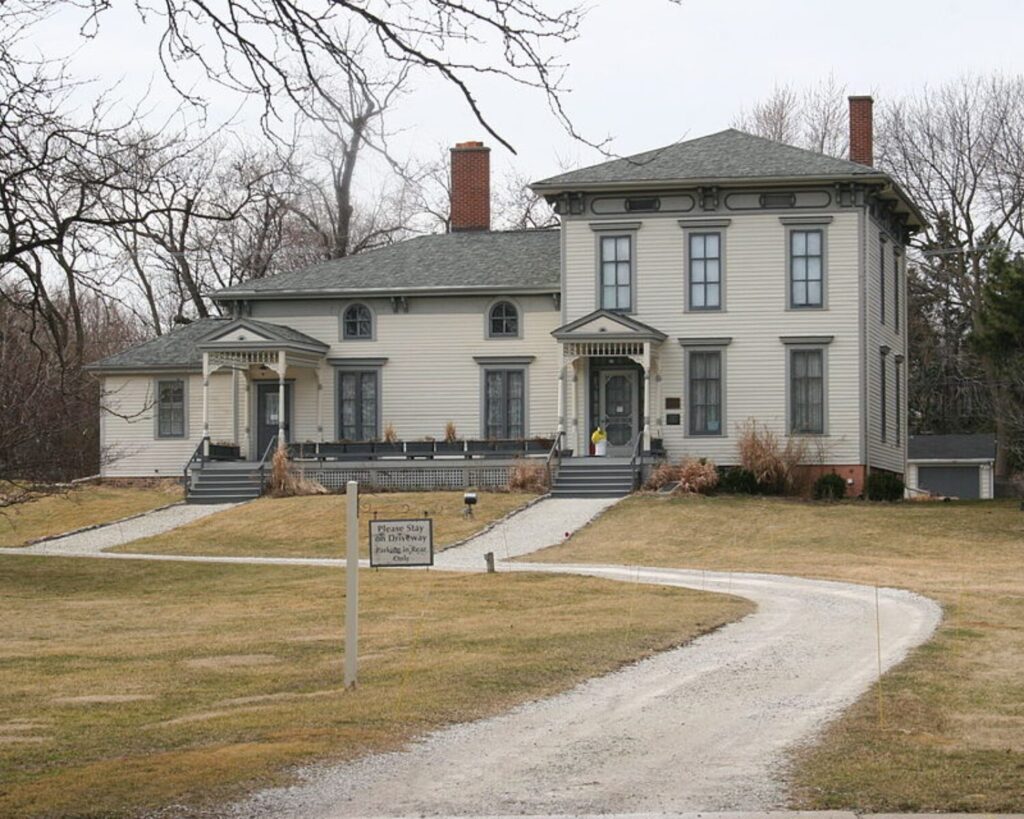 Historic Noble–Seymour–Crippen House with pale siding, front porches, brick chimneys, and a curved gravel driveway framed by leafless trees.