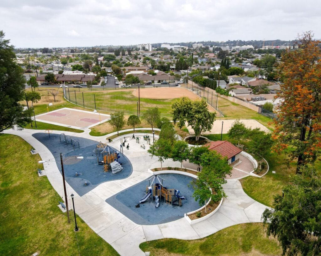 Aerial view of a suburban park with a blue playground, basketball court, baseball field, and surrounding houses.