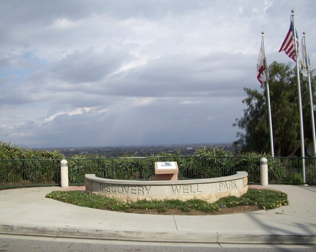 Discovery Well Park overlook with stone “Discovery Well Park” sign, flags, cloudy sky, greenery, and distant city view.