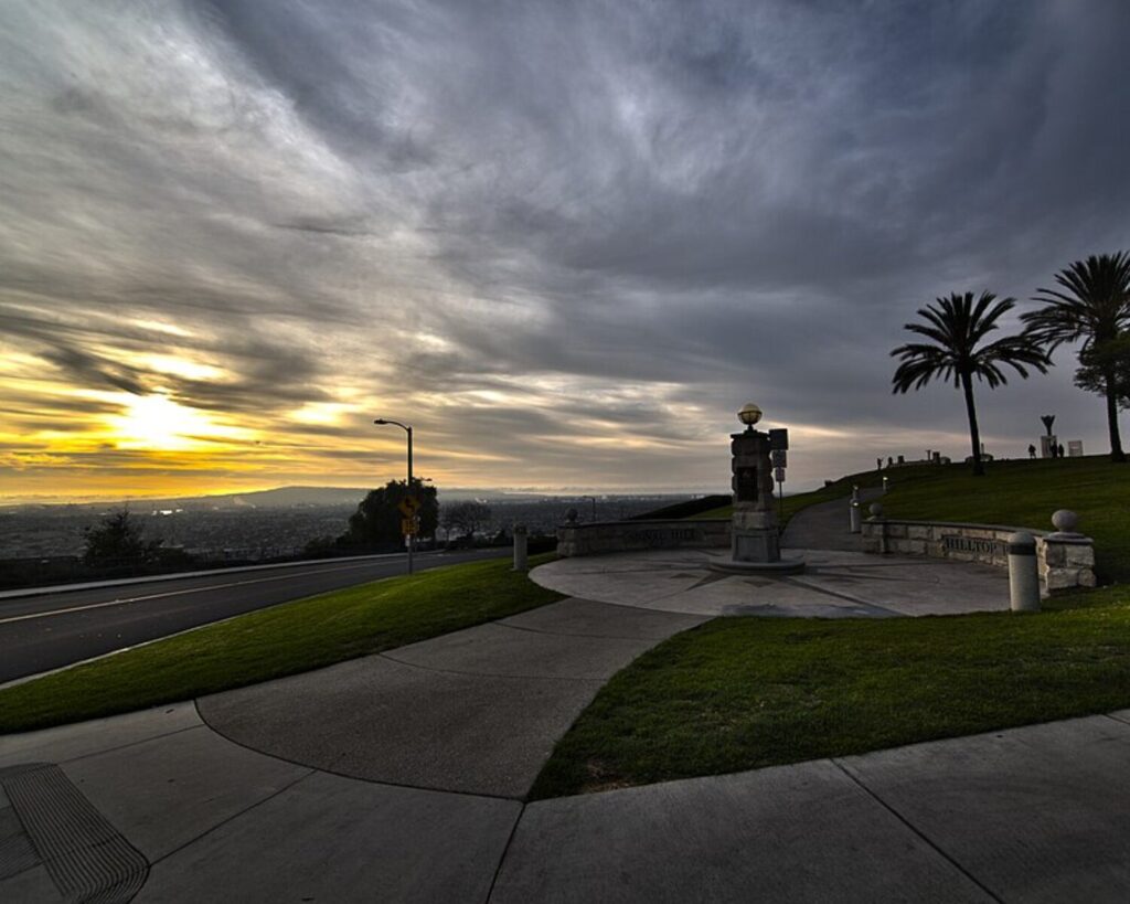 Dramatic sunset view from Signal Hill Hilltop Park with cloudy sky, palm trees, road, green lawn, and distant city skyline.