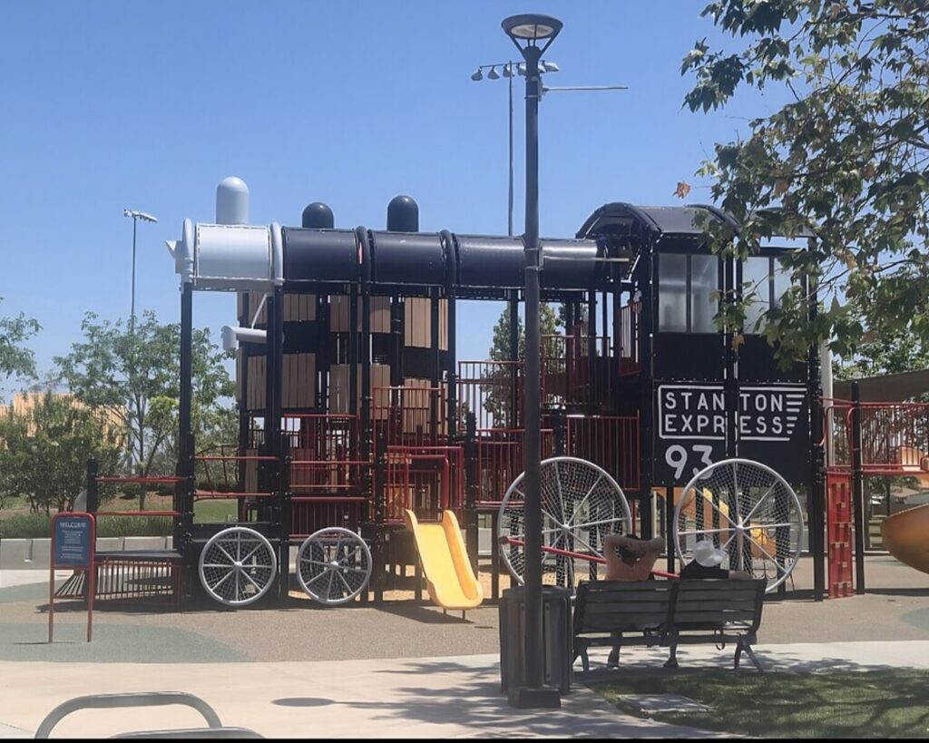 Central Park Stanton playground with train-themed play structure, slides, bench, trees, and clear blue sky.