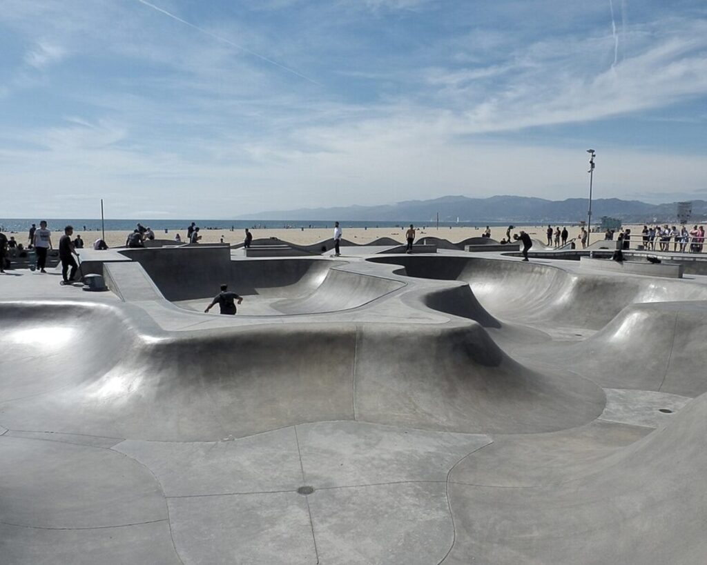Seaside skatepark with smooth concrete bowls and skaters, ocean and mountains in the background.