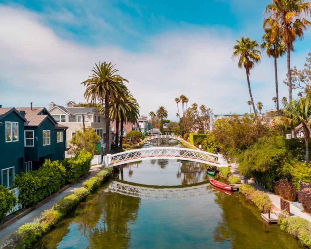 Venice Beach canal lined with homes and palm trees, featuring a white pedestrian bridge and red canoe.
