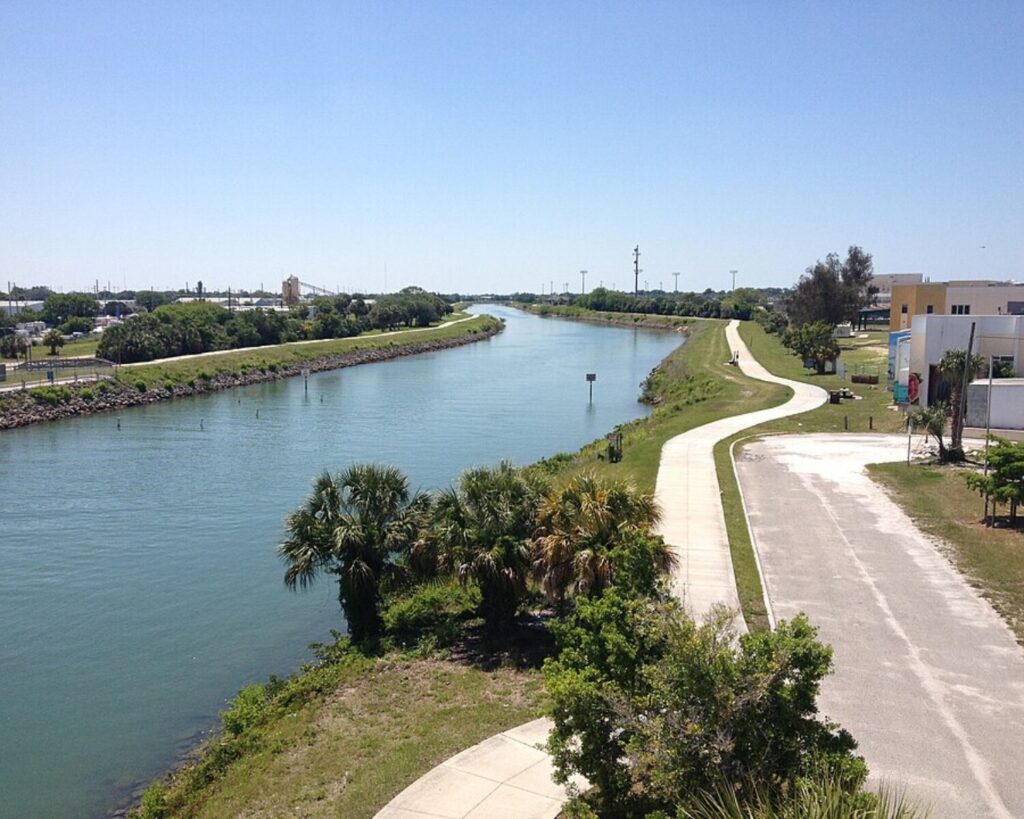 Wide canal with a curved path along the right, trees and grassy banks under a clear blue sky