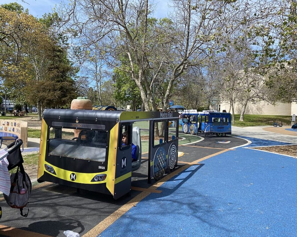 Bus-themed playground equipment at Westwood Recreation Center with children, trees, and blue play surface.