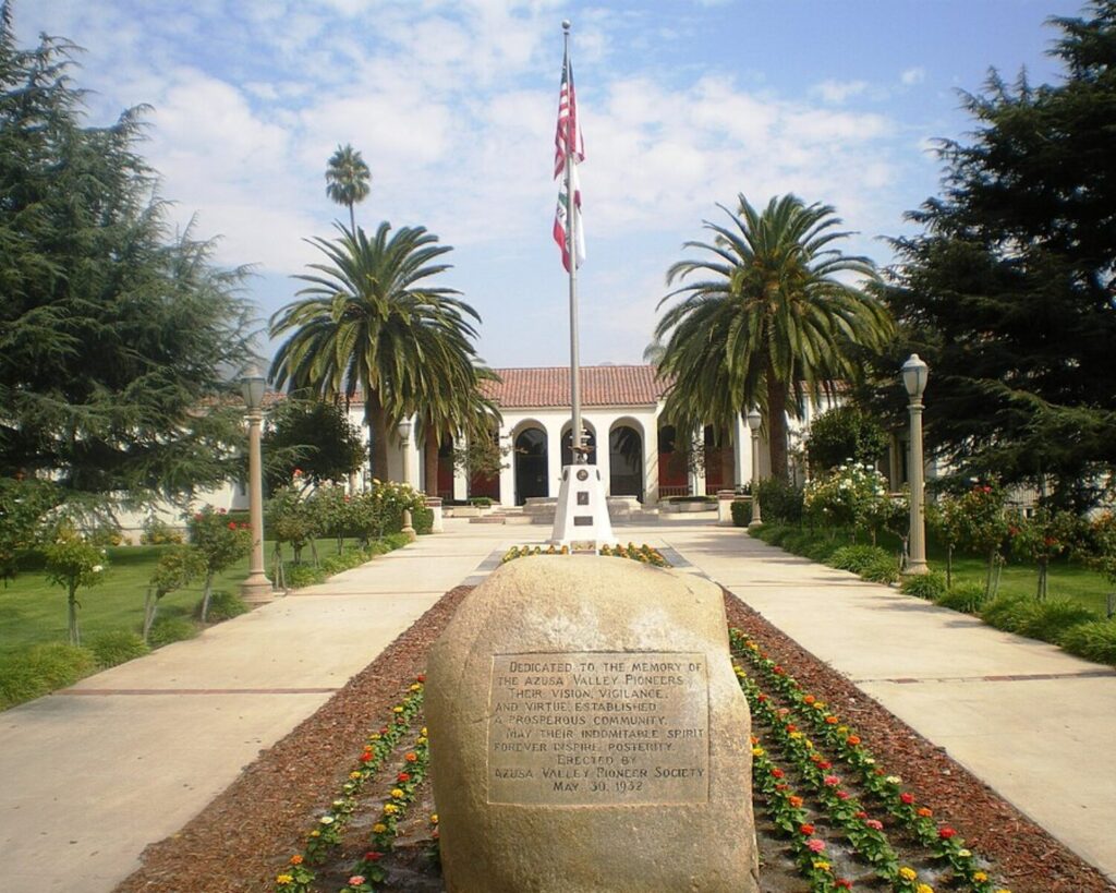 Azusa City Hall framed by palm trees and landscaped gardens, with flagpoles and a memorial stone in front.
