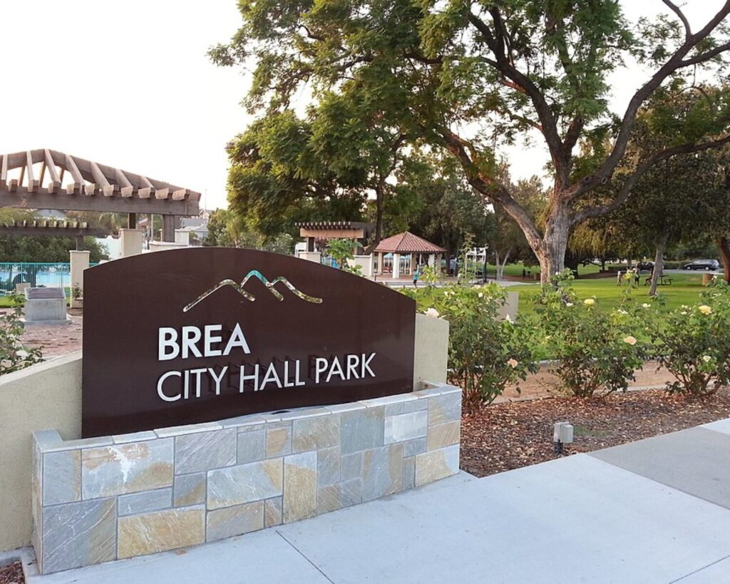 Brea City Hall Park entrance sign surrounded by landscaped gardens, trees, walkways, and park structures.