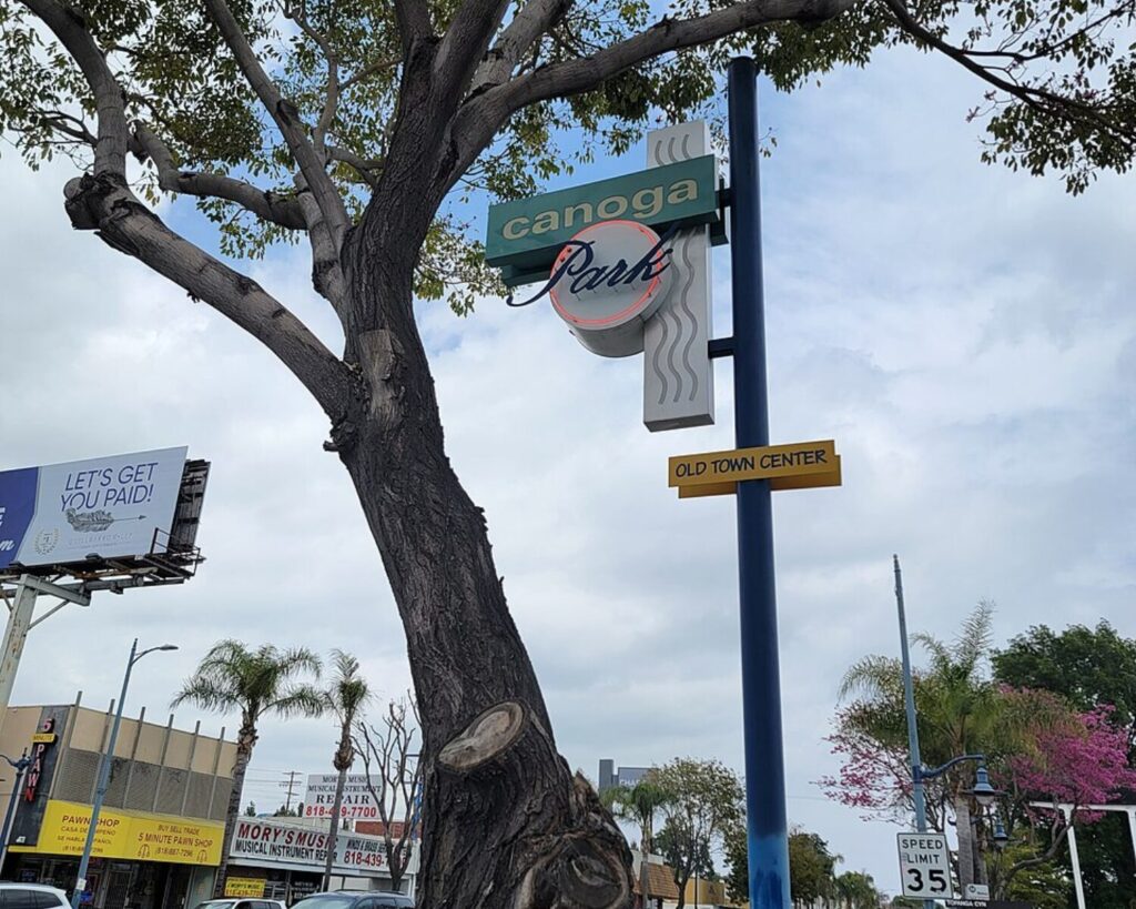 Canoga Park Old Town Center sign beside a large tree, with storefronts and a commercial street in the background.