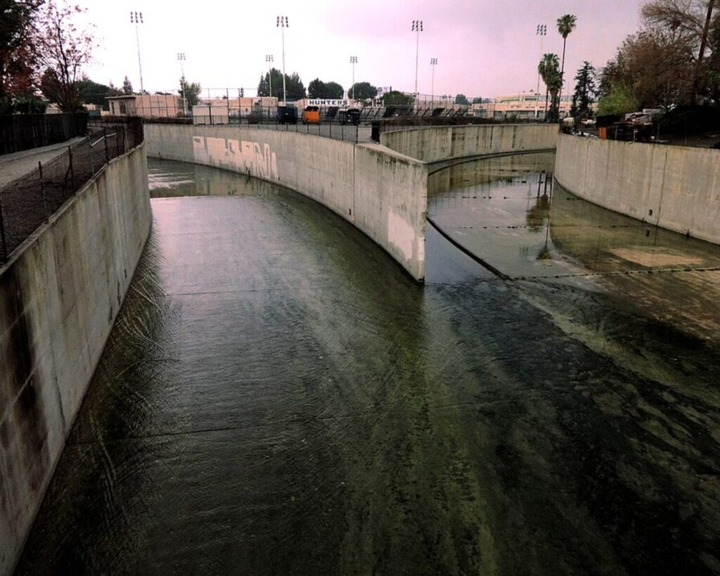 Concrete canal with curved walls and flowing water, fences along the sides and distant flood-light towers in a parking area beyond the top edge.