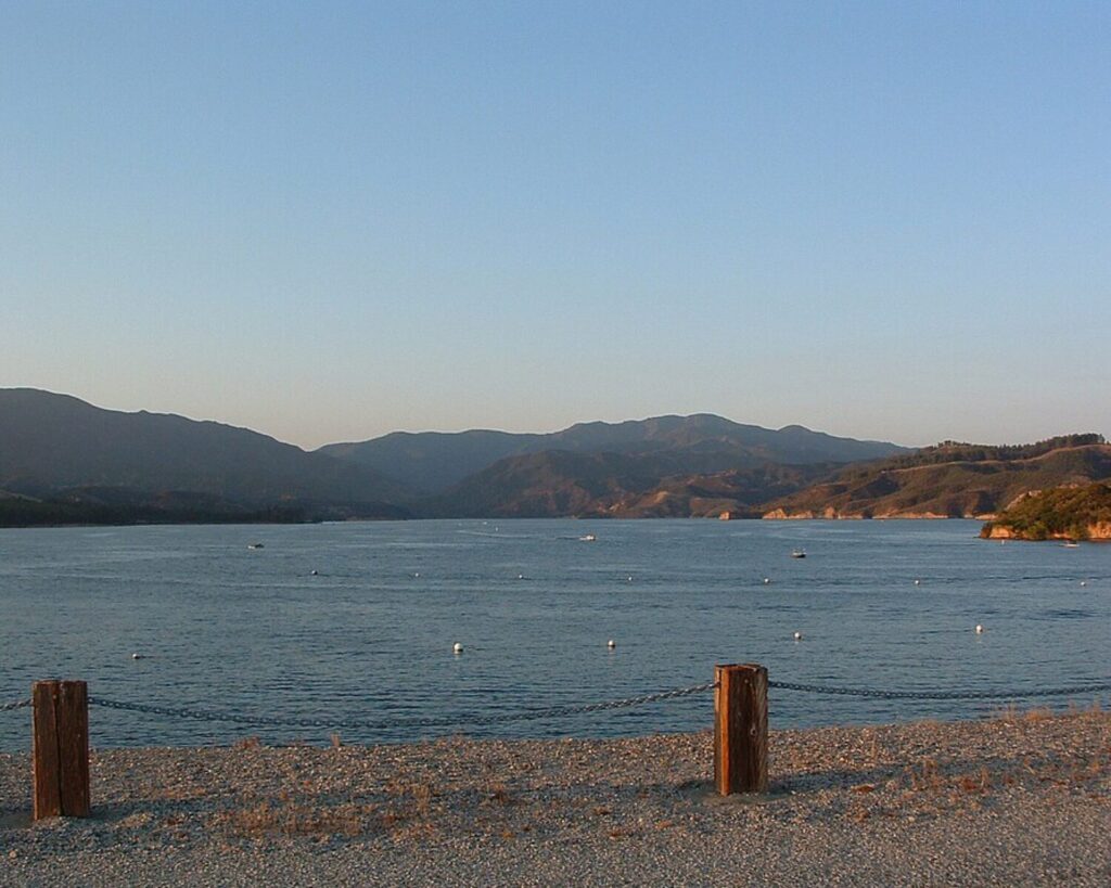 Castaic Lake surrounded by rolling hills, with calm blue water, scattered boats, and a rocky shoreline.