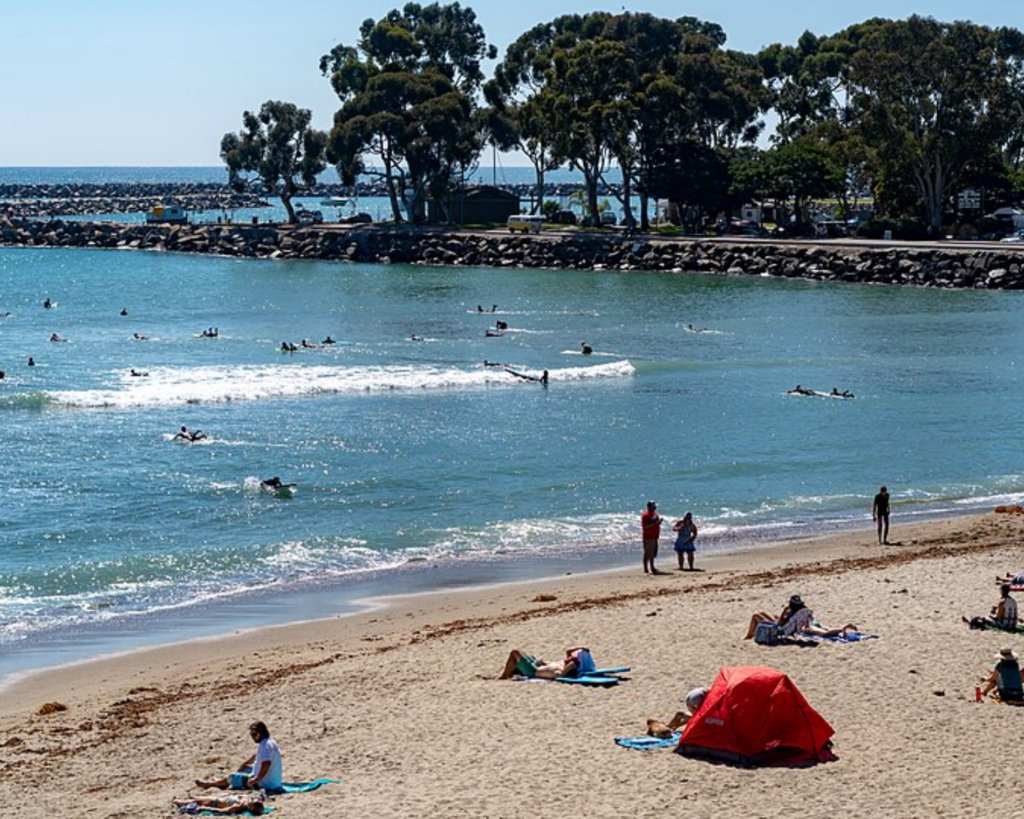 Sunlit beach scene with people swimming and surfing in turquoise water and a red tent on the sand in the foreground.