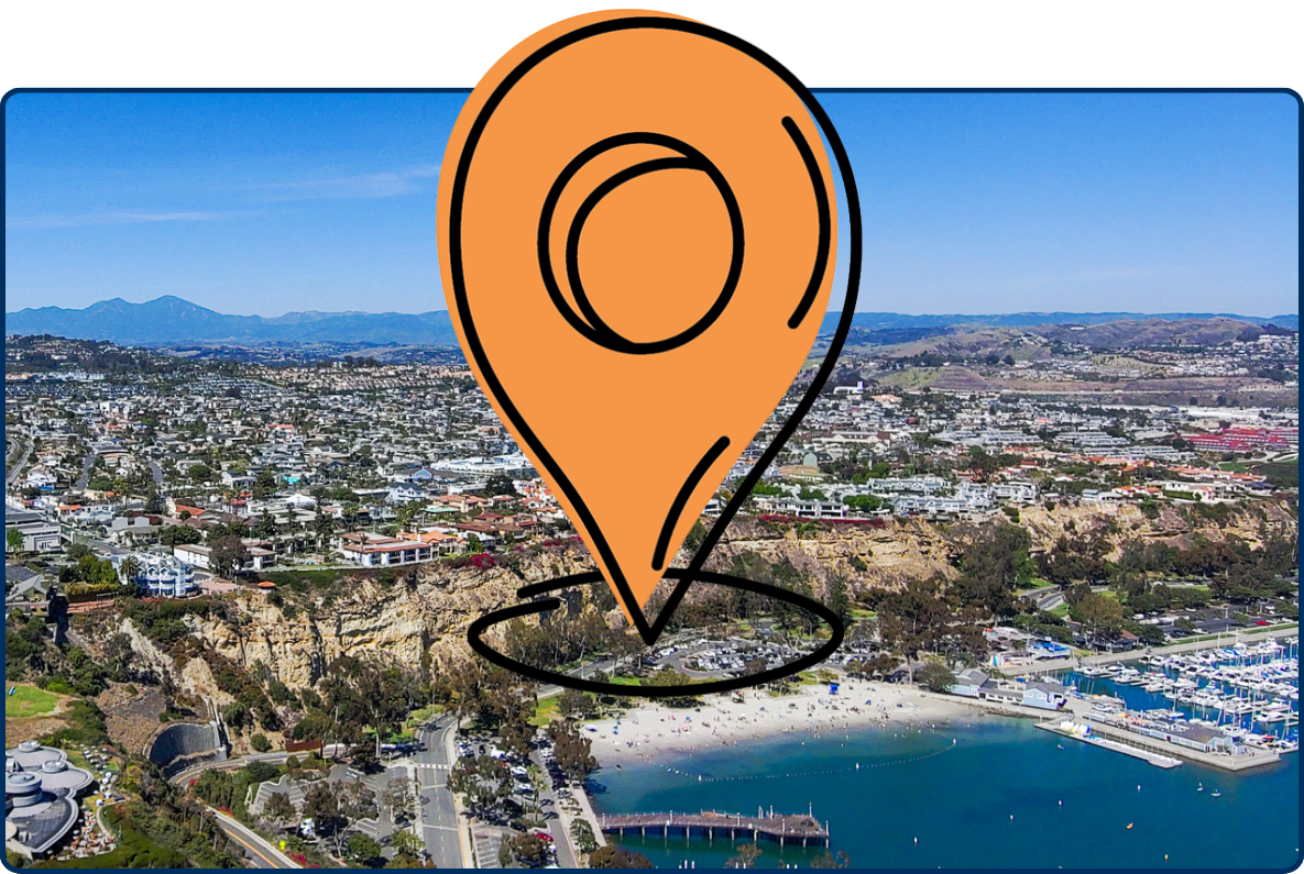 Aerial view of Dana Point, California, with the harbor, coastal roads, hillside neighborhoods, and mountains under a clear blue sky.