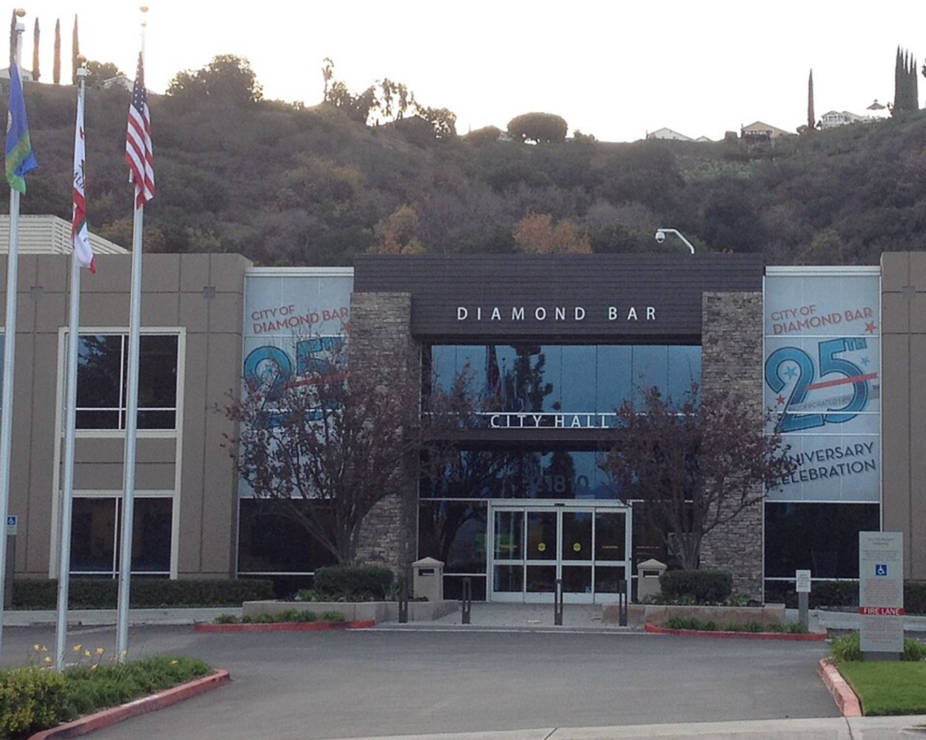 Front view of Diamond Bar City Hall with flagpoles, landscaped grounds, and hills rising behind the building.