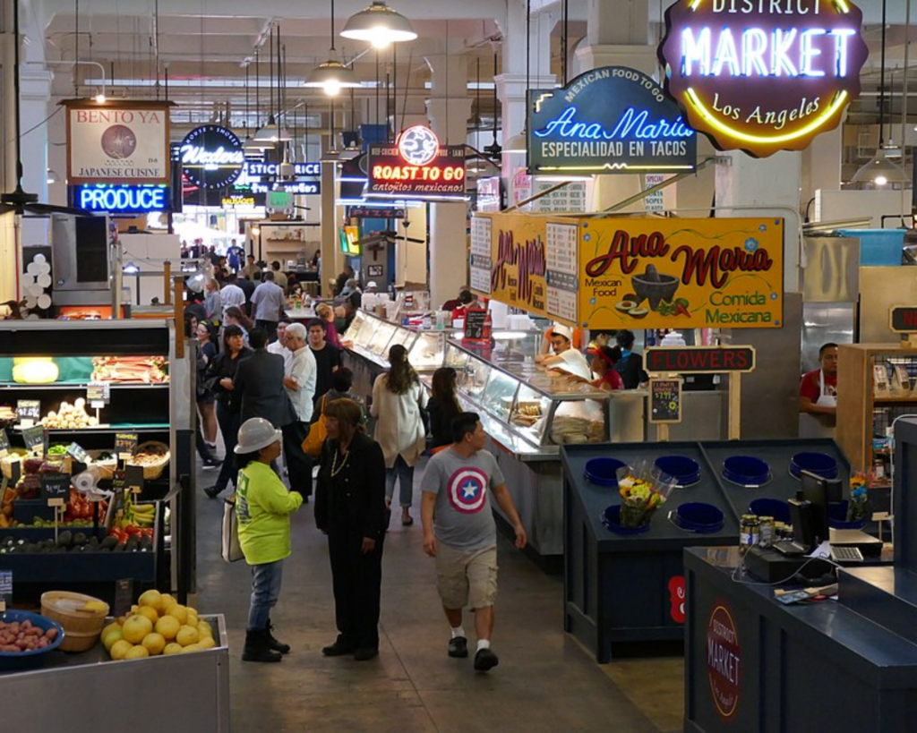 Busy interior of Grand Central Market in Los Angeles with food stalls, produce stands, neon signs, and shoppers.