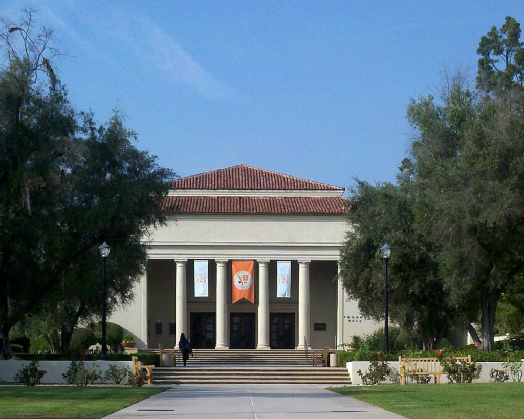 Neoclassical campus building with red-tile roof, tall columns, banners, and broad stairs on a sunny day