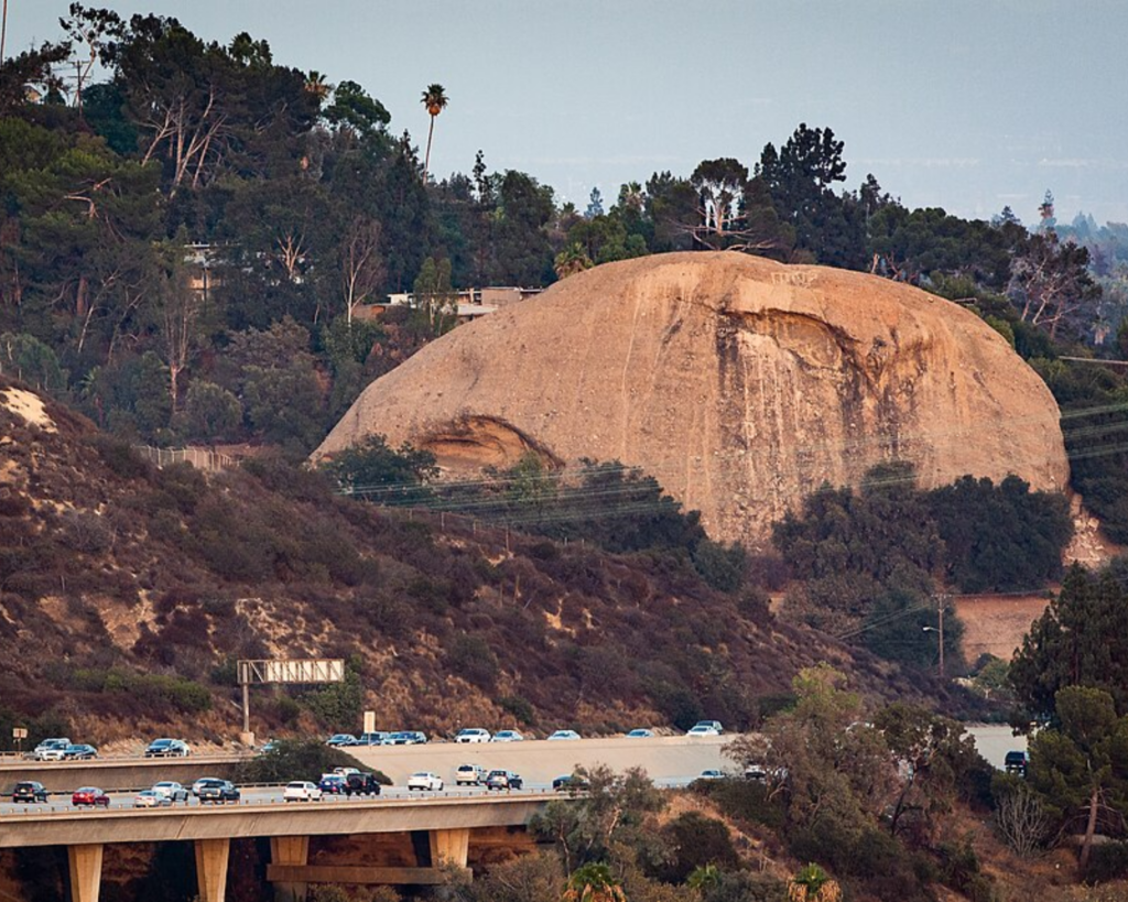 Large rounded rock formation on a hillside with a highway and cars in the foreground, surrounded by trees and shrubland