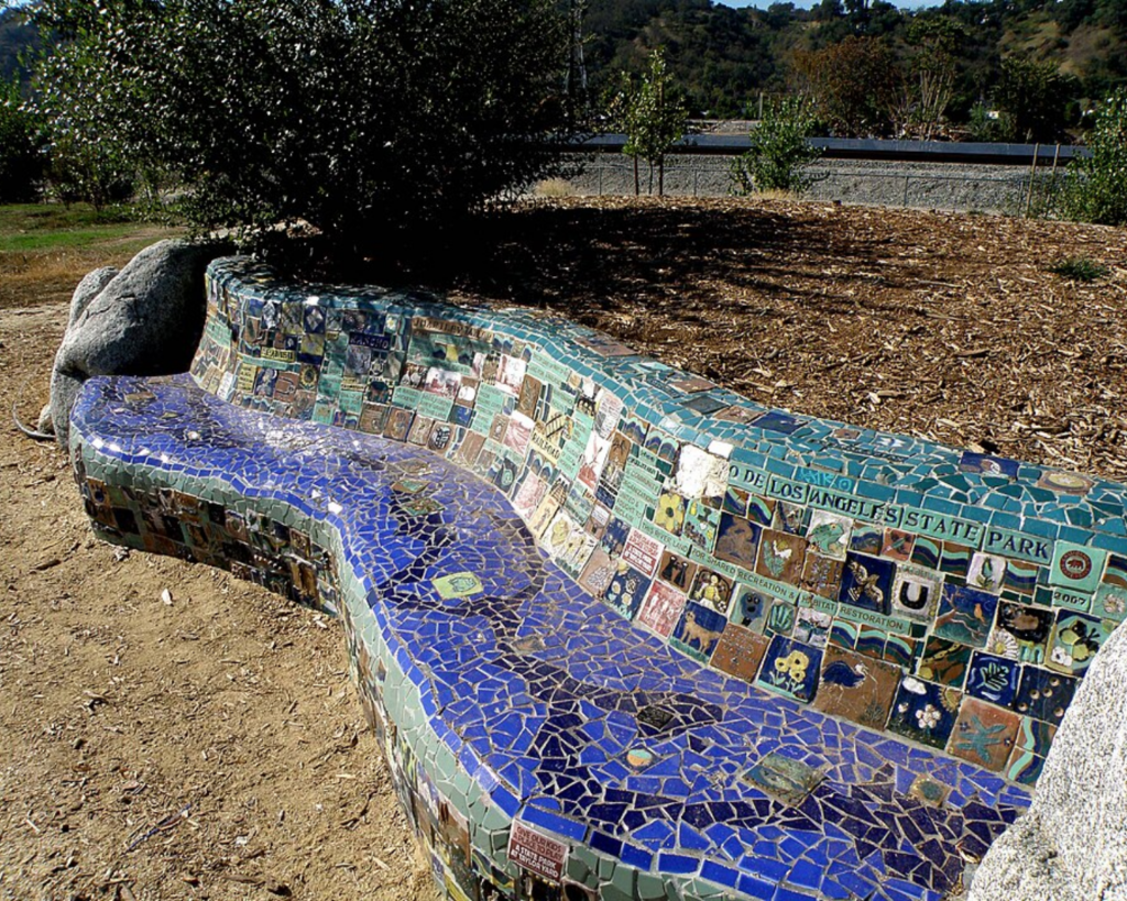Curved mosaic bench in a park, covered in small colorful tiles with shades of blue and teal, beside a dirt area and shrubs.