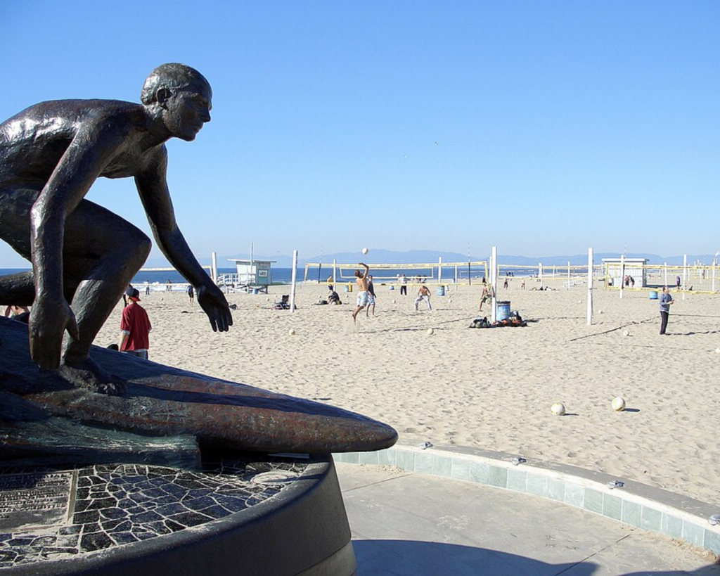 Bronze crouching statue on a circular pedestal at the beach, with volleyball nets in the background, sunny day