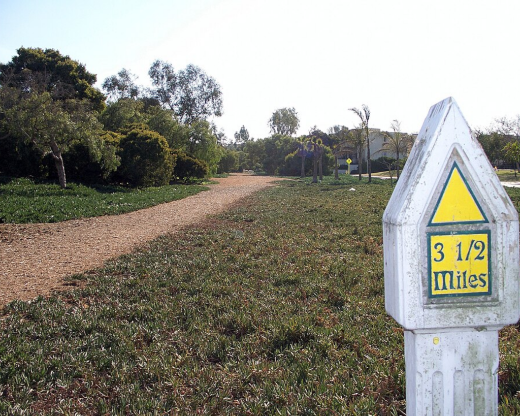 White milestone post with a yellow triangle and a blue-bordered panel reading '3 1/2 Miles' beside a gravel park path; trees and a sunny sky in the background.