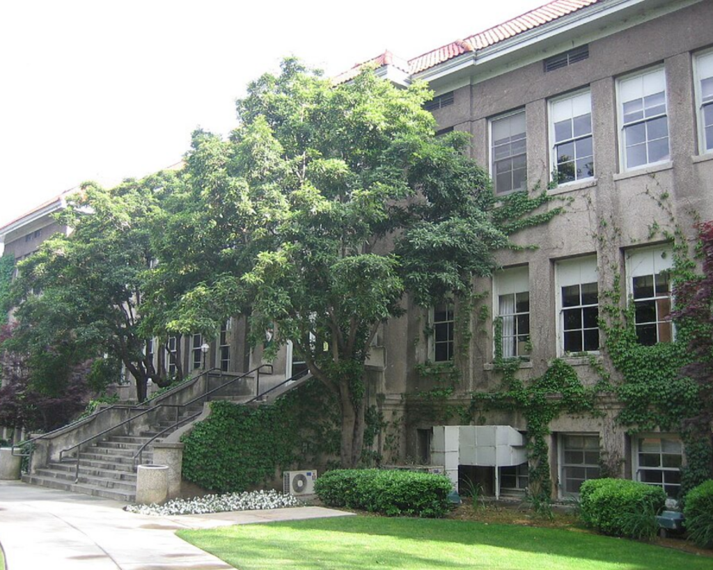 Campus-style building with ivy-clad walls, large windows, and a set of concrete stairs leading to the entrance, surrounded by trees and a manicured lawn.