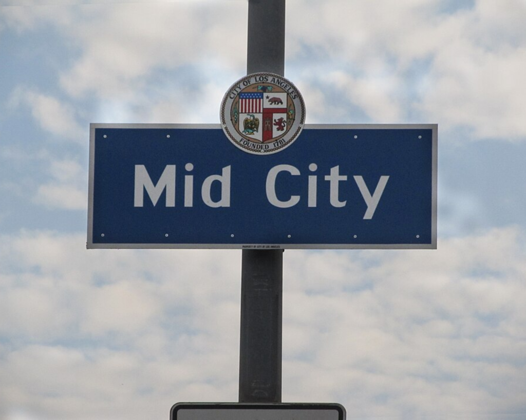 Mid-City neighborhood sign in Los Angeles mounted on a pole beneath the city seal against a cloudy sky.