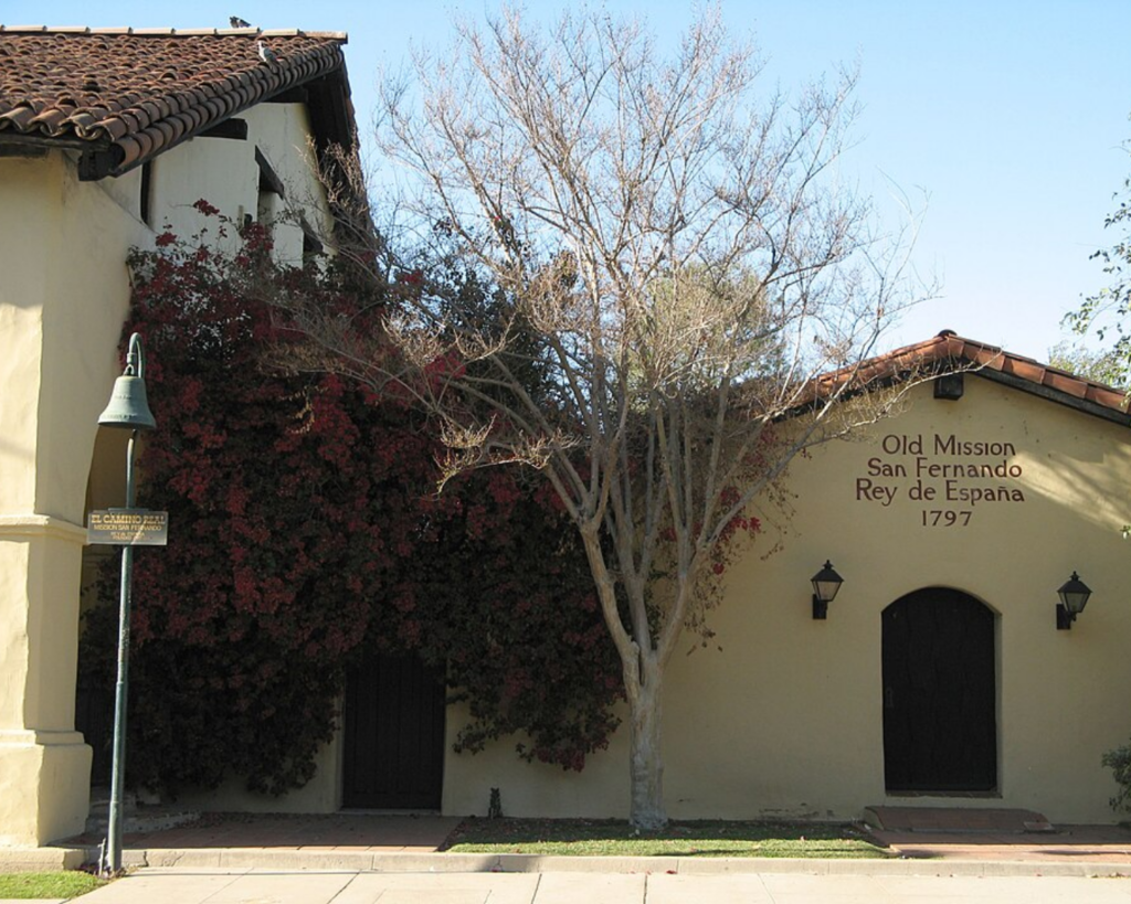 Old Mission San Fernando Rey de España in Los Angeles, featuring historic adobe-style buildings, tiled roofs, a bell, and surrounding trees.