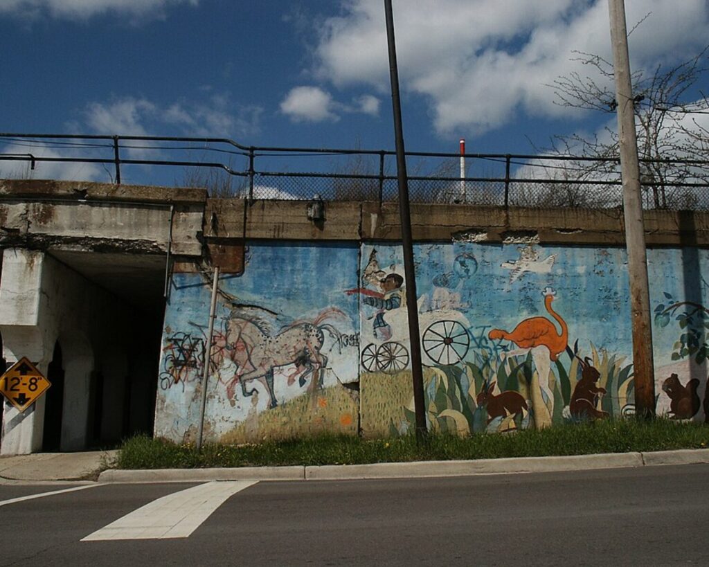 Weathered mural beneath a railway overpass in Chicago’s Pilsen neighborhood, depicting horses, a wagon, birds, and other animals.