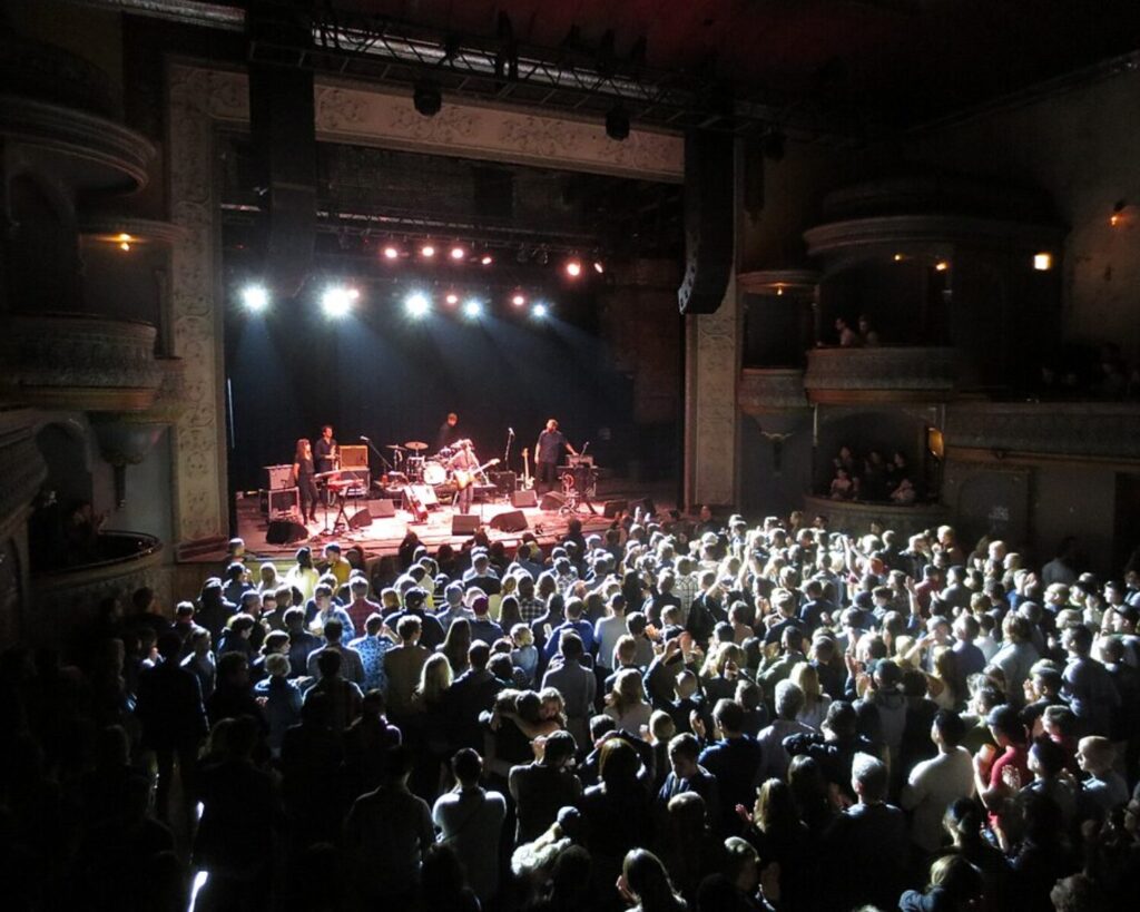 Concert hall scene with a band on stage performing to a large audience under bright stage lights in a decorated theater.