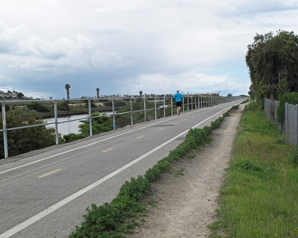 Ballona Creek Bike Path in Los Angeles, with a paved cycling trail, creekside vegetation, fencing, and a person walking under a cloudy sky.