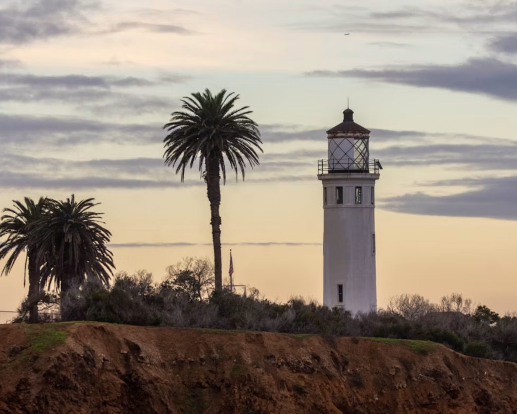 White lighthouse surrounded by tall palm trees on a coastal cliff beneath a softly clouded sunset sky.