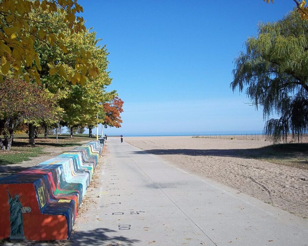 Path through Loyola Park leading to a sandy Lake Michigan beach, lined with autumn trees and colorful painted barriers.