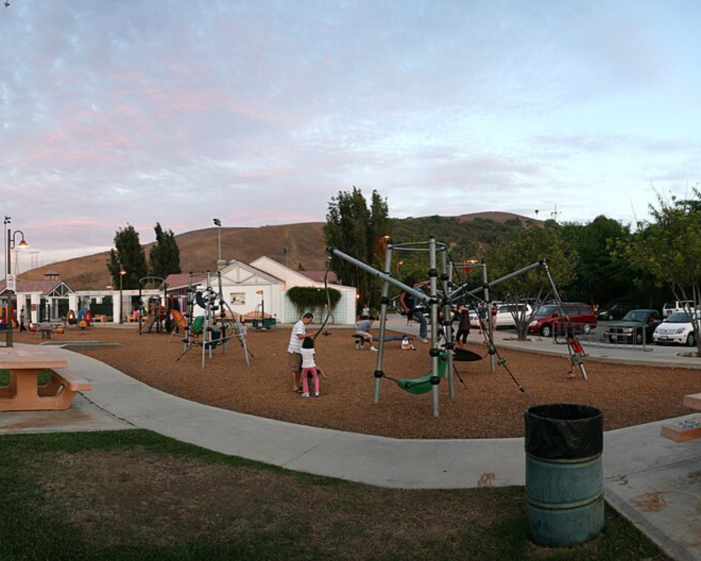 Pathfinder Park playground with climbing structures, children playing, picnic tables, parked cars, and rolling hills in the background.