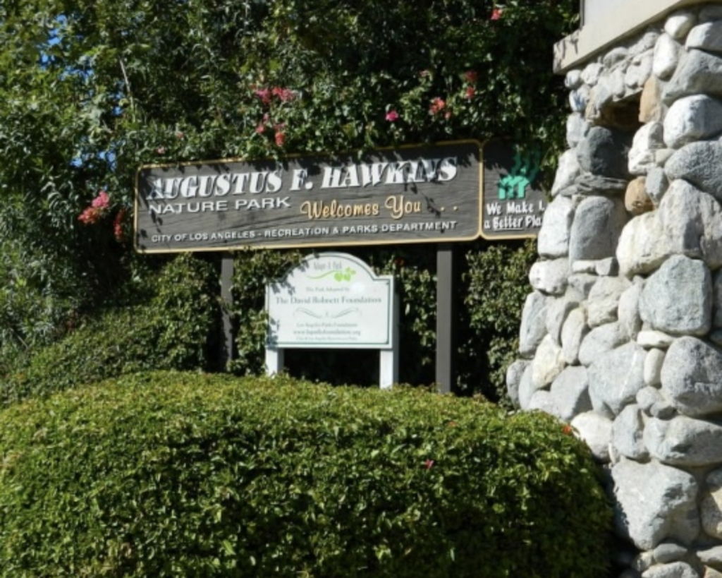 Augustus F. Hawkins Nature Park entrance sign in Los Angeles, surrounded by dense greenery beside a stone wall.