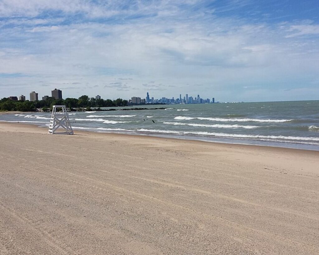 Wide sandy Rainbow Park Beach beside Lake Michigan, with waves, a lifeguard chair, and the Chicago skyline in the distance.