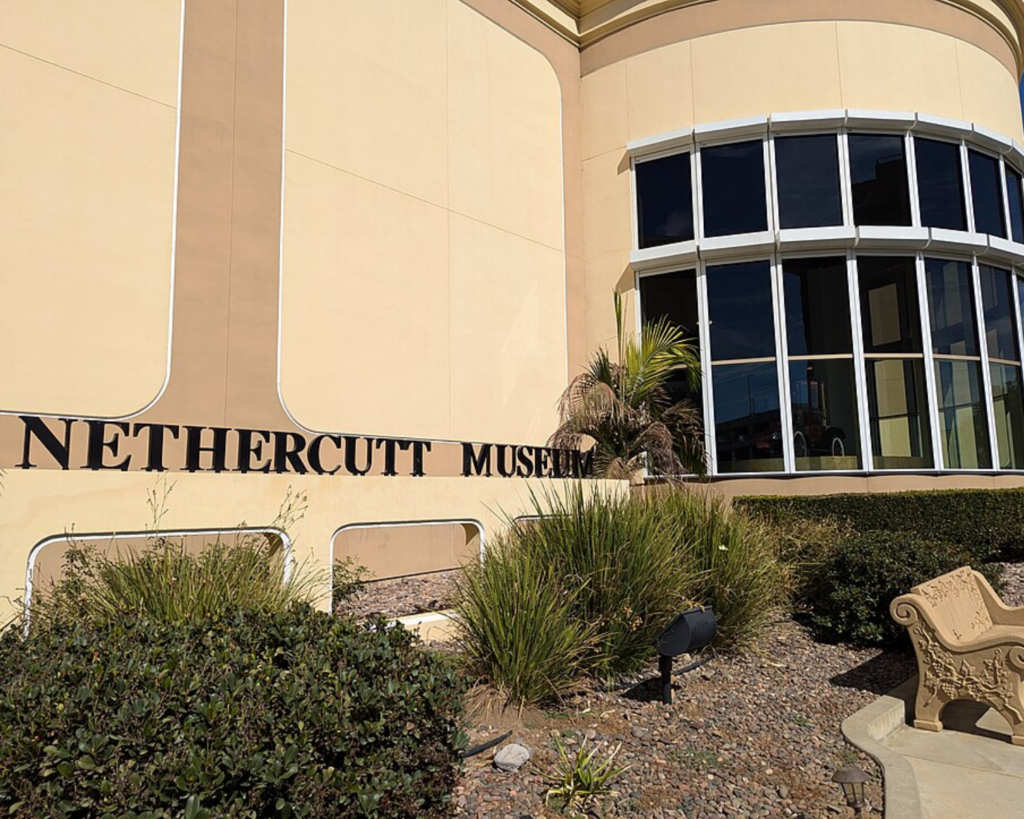 Nethercutt Museum exterior in Sylmar, California, featuring a beige facade, curved glass windows, landscaped grounds, and the museum name displayed on the building.